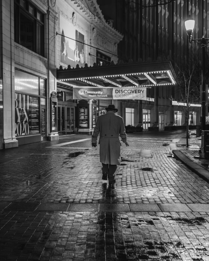 Street photo of a man in a long coat and hat walking on wet streets.