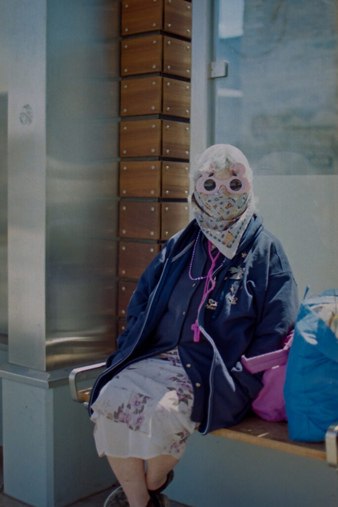 The masked woman sitting at a bus stop wearing a mask and pink glasses.